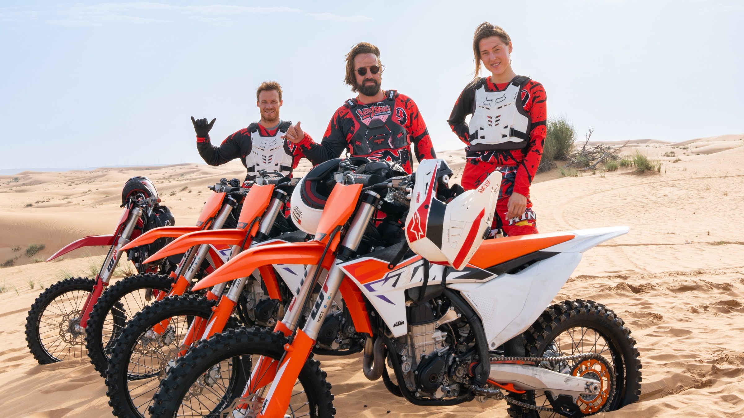 Three people in motocross gear stand by dirt bikes on sand dunes under a clear sky.