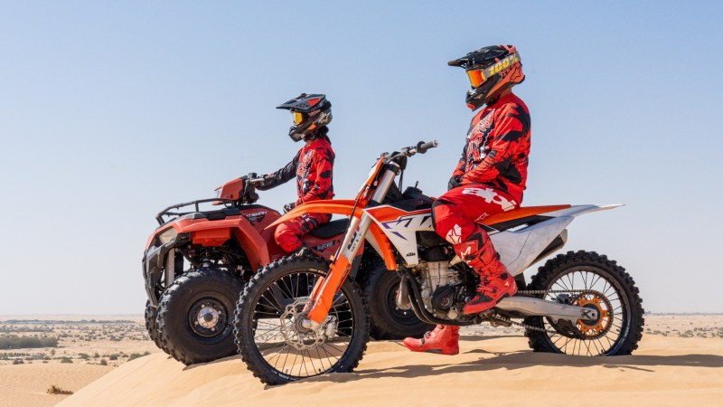 Two people in red gear on an ATV and dirt bike in a desert landscape.