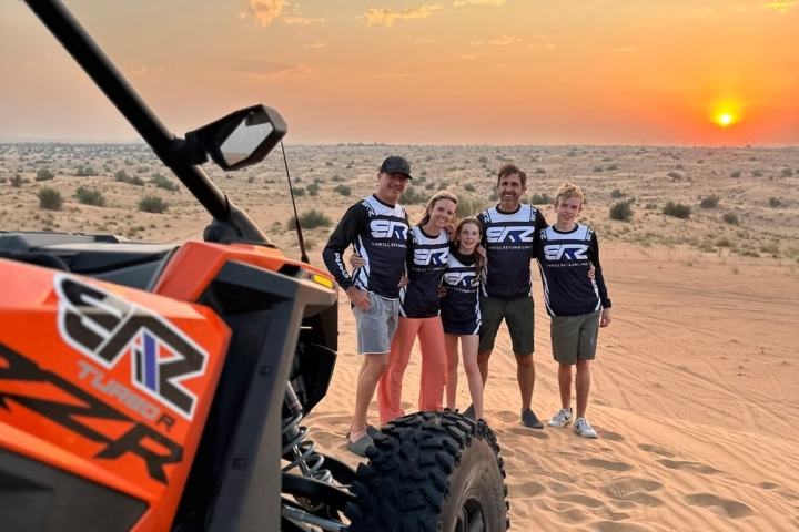 Group of five posing with dune buggy in desert at sunset.