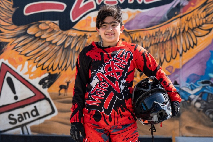 Person in red motocross gear holding a helmet, standing before a colorful 'Moto Zone' mural.