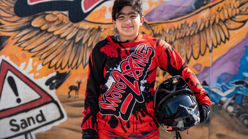 Person in red motocross gear holding a helmet, standing before a colorful 'Moto Zone' mural.