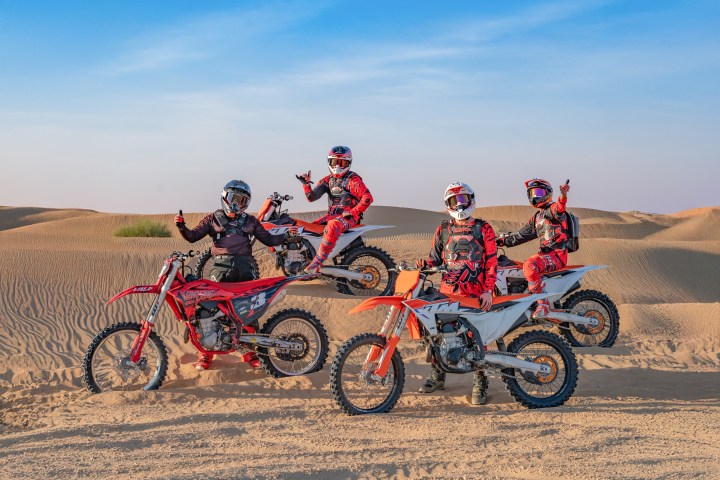 Four motocross riders in vibrant gear pose in desert dunes at sunset.