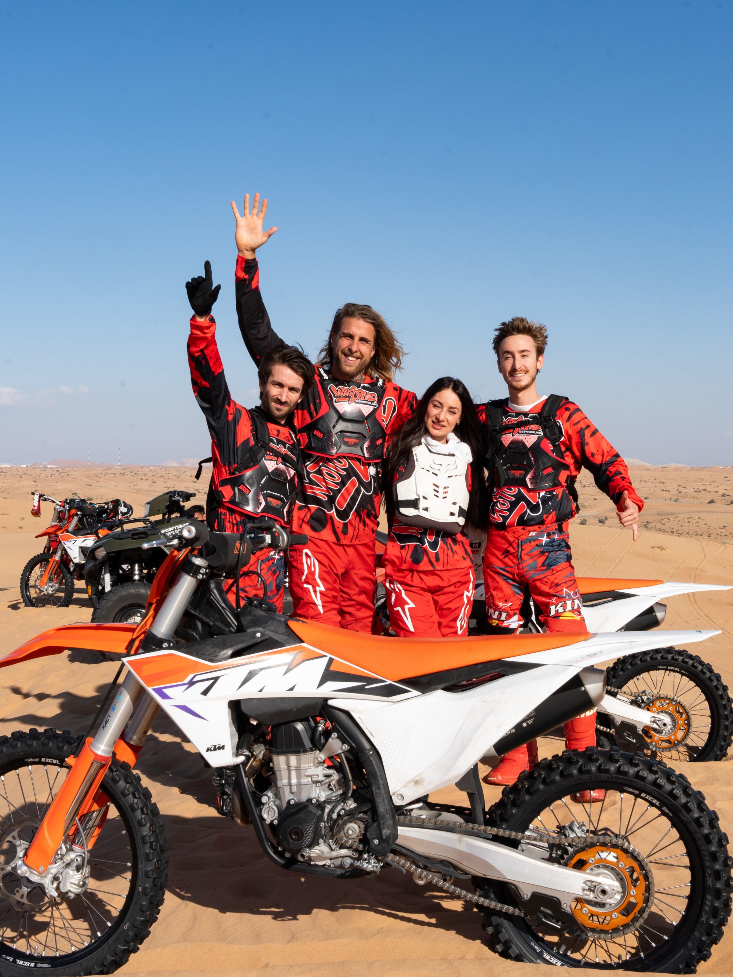 Four people in red gear standing beside dirt bikes in a desert landscape.