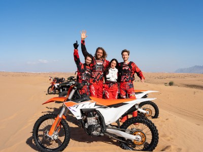 Four people in red gear standing beside dirt bikes in a desert landscape.
