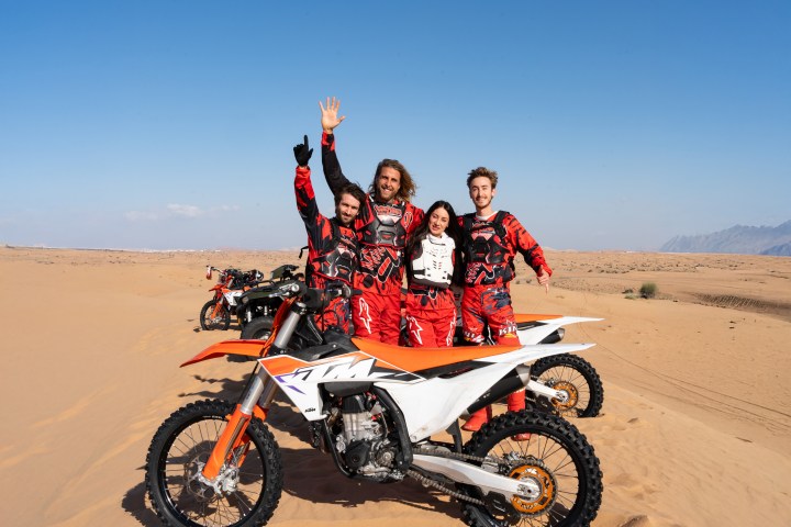 Four people in red gear standing beside dirt bikes in a desert landscape.
