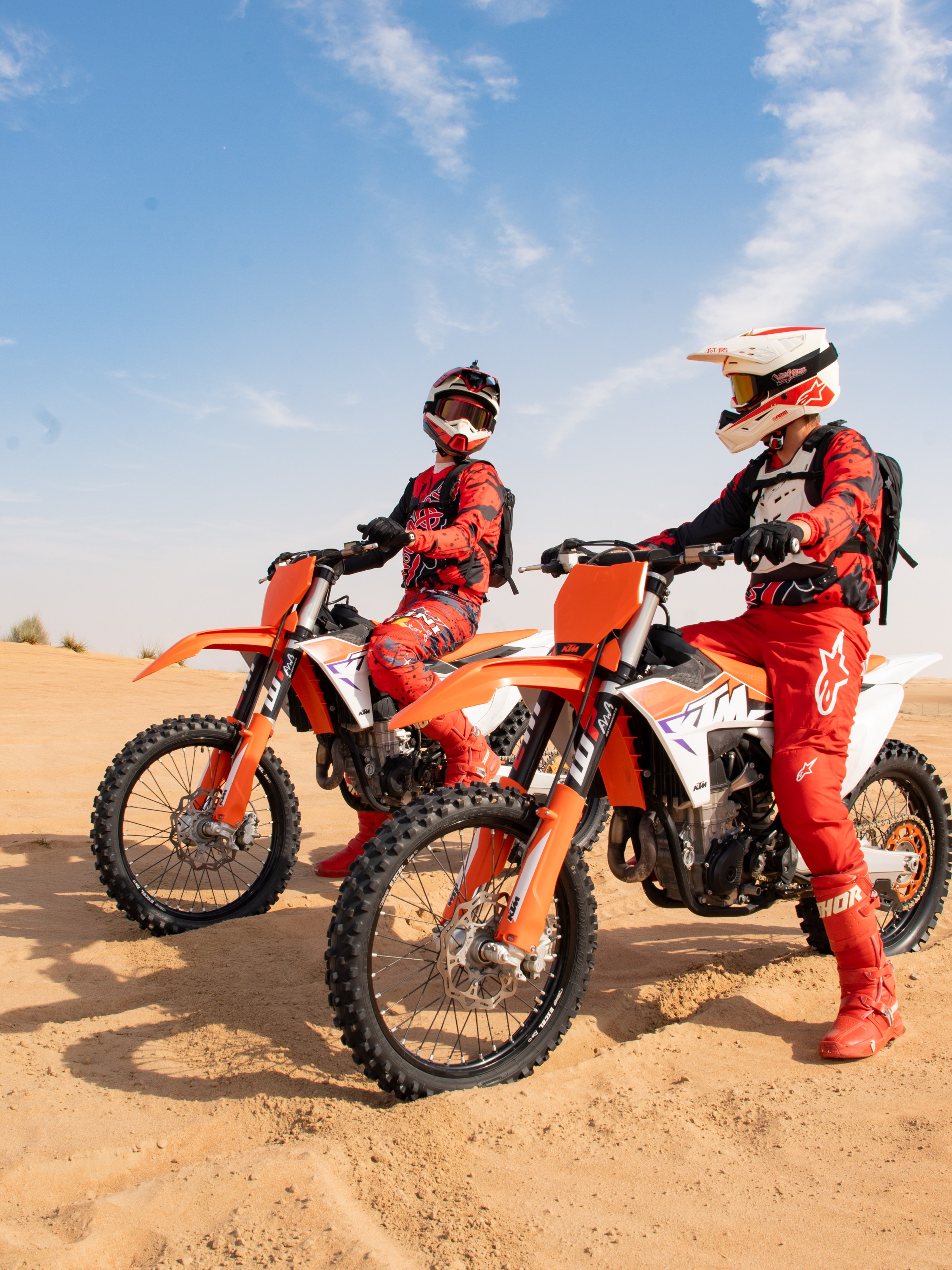 Two motocross riders in red gear on orange dirt bikes in a sandy desert.