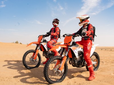 Two motocross riders in red gear on orange dirt bikes in a sandy desert.