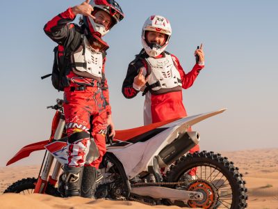 Two motocross riders in red gear standing beside a bike in a desert landscape.