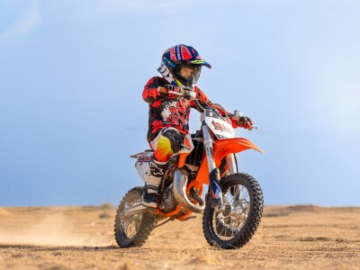 Child in motocross gear riding a dirt bike on sandy terrain.